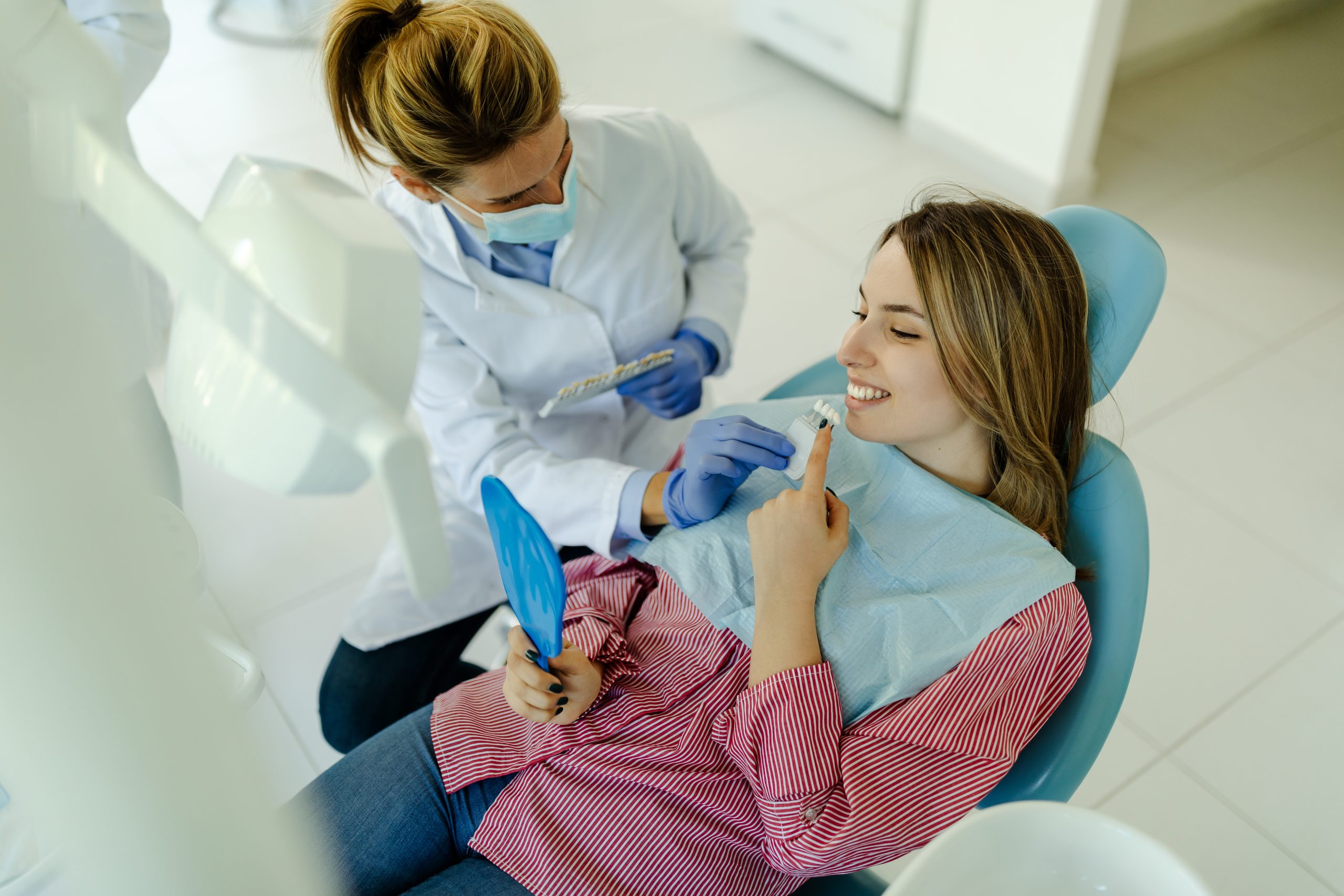 Young Woman Sitting In Dental Chair And Selecting Best Color Of Implants Using Teeth Samples.